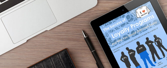 A workspace featuring a laptop, an iPad displaying a document titled "The Success of Loyalty Programs," and a pen.