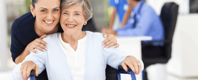 A smiling older woman in a wheelchair, with a younger woman standing behind her.