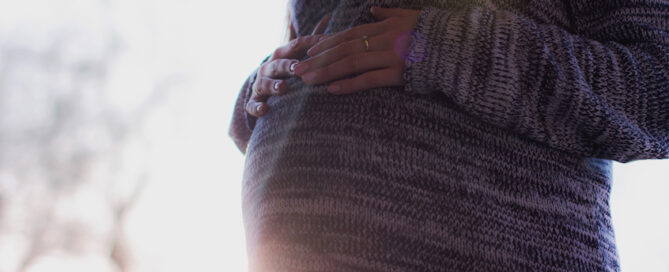 Woman standing, gently holding her pregnant belly with both hands.