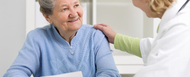 A healthcare professional is sitting across an elderly woman and gently resting a hand on the woman's shoulder.