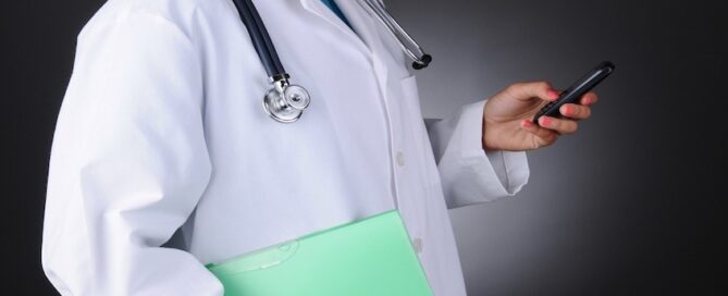Doctor with a stethoscope around their neck is holding a smartphone in one hand and a green folder in the other.