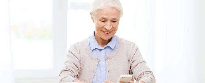 A smiling elderly woman is sitting at a table, looking at her smartphone.