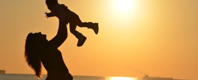A silhouette of a woman lifting a child above her head against a bright sunset backdrop.