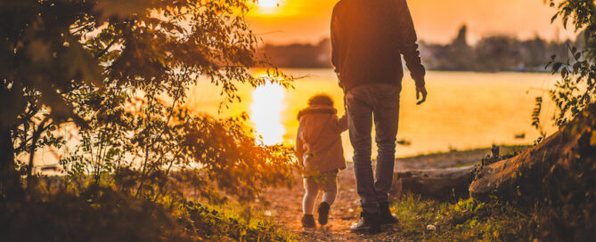 A silhouette of a man and a child walking hand in hand along a path by the water during sunset.
