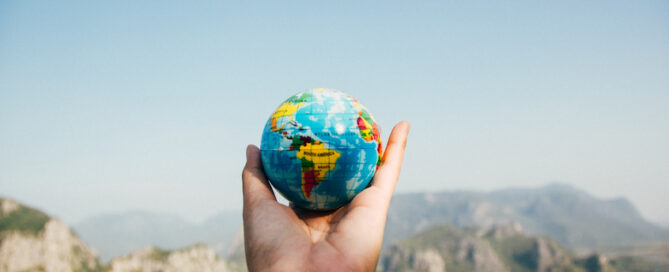 A hand is holding a small globe against a backdrop of mountains and clear blue sky.
