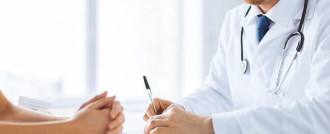 Male doctor is sitting at a desk, focused on writing notes while a patient's hands can be seen clasped together across the table.