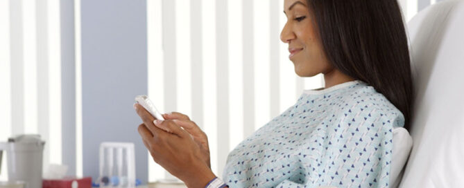Woman is sitting in a hospital bed, looking at her smartphone.
