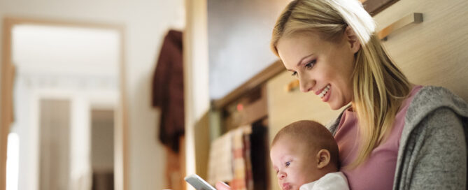 Woman is sitting on the floor, holding a baby in her lap, as she looks at her smartphone.