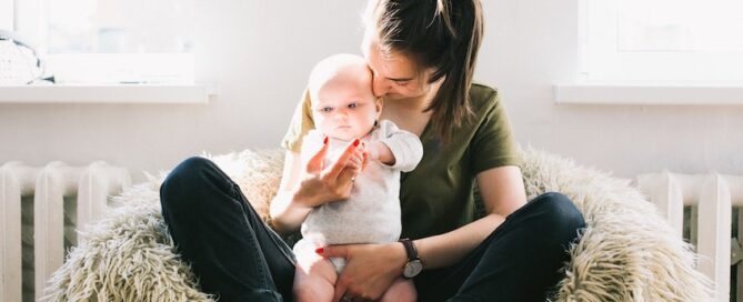 A woman sitting on a cozy, fluffy chair holding a baby gently kisses the baby's forehead.