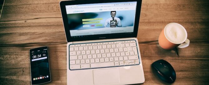 A laptop placed on a wooden surface, with a smartphone, a computer mouse and a beverage beside it.