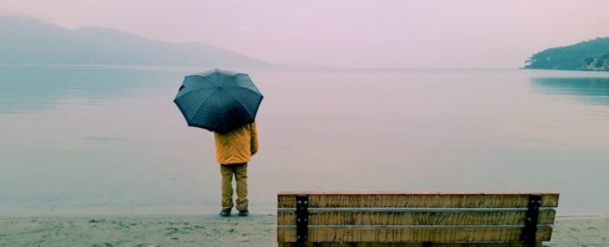 A person standing on a beach, holding an umbrella, facing a calm body of water.