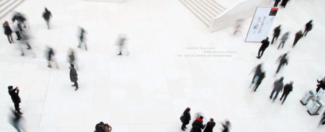 An image depicts a spacious, brightly lit interior with white walls and flooring. People are moving throughout the space in various directions, creating a sense of dynamic activity.