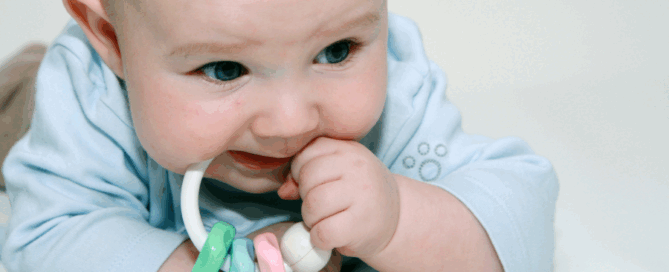 Baby is lying on their stomach, holding a colorful teething toy with their right hand and bringing it to their mouth.