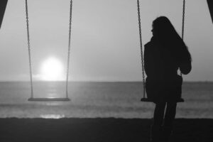 A silhouette of a person sitting on a swing, looking out towards the horizon where the sun is setting over the ocean.