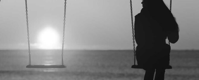 A silhouette of a person sitting on a swing, looking out towards the horizon where the sun is setting over the ocean.