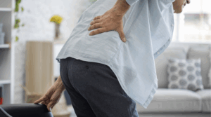 Person is seen from behind, standing and leaning slightly forward with one hand on their lower back, appears to be experiencing discomfort or pain.