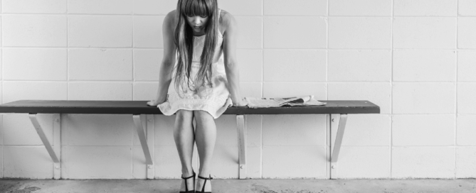 Girl in a white dress sits on a wooden bench, with her head down, looking dejected.