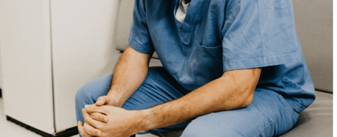 Healthcare worker seated in a clinical setting, appears to be in a moment of contemplation or stress.