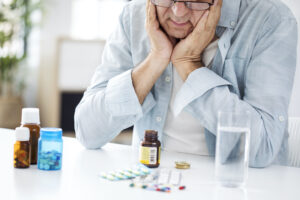 Old man sitting at a table with a concerned expression, holding his head in his hands, with various types of medicine bottles.
