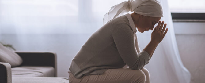 Woman sitting on a couch with their head bowed and hands clasped together in front of their face, wearing a headscarf and appear contemplative or distressed.