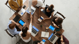 Group of people engaged in a business meeting or discussion around a large wooden table, with laptops and electronic devices in front of them.
