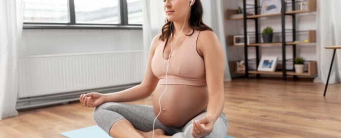 Happy pregnant woman with earphones and smartphone listening to music and meditating at home.