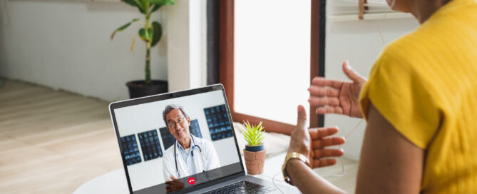 Woman sitting at a table engaging in a video call with a male doctor on her laptop.