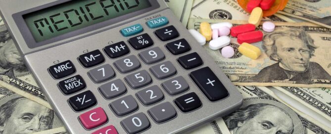 Gray calculator displaying the word "MEDICAID," placed on a bed of U.S. dollar bills with prescription bottle and various colorful pills.