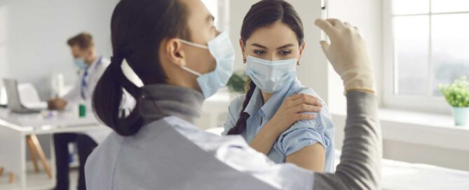 Healthcare professional preparing a syringe while standing near a woman who is receiving a vaccination.