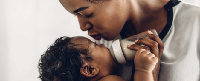 Mother tenderly leaning in to kiss her baby, who is drinking from a bottle.