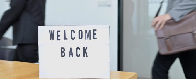 Sign that says "WELCOME BACK" is placed on a wooden table, with people walking in the background.
