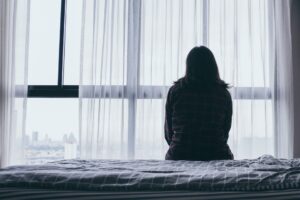 Woman sitting on a bed, facing a large window covered with sheer curtains.