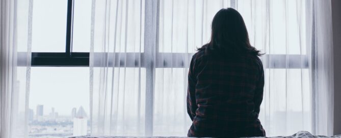 Woman sitting on a bed, facing a large window covered with sheer curtains.