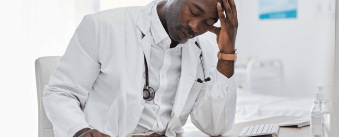 Male doctor siitting at a desk, appears to be stressed or overwhelmed, with his hand on his forehead.