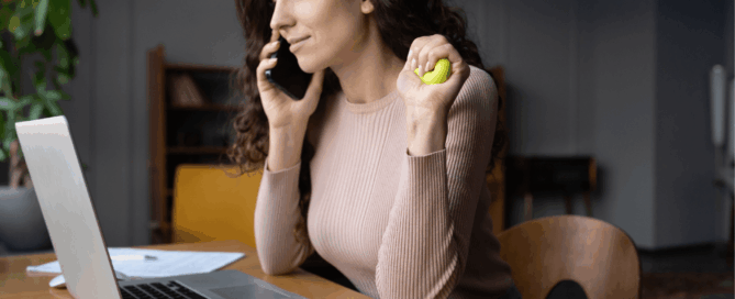 Woman in front of a laptop answering a call on her phone while holding a stress ball.