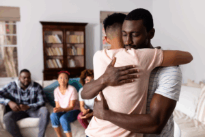 Two men embracing, with several people sitting on a couch.