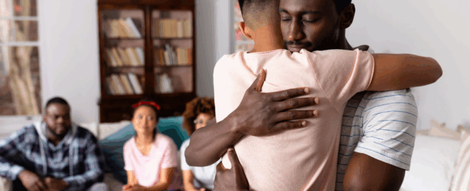 Two men embracing, with several people sitting on a couch.