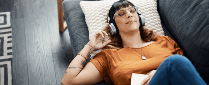 Woman is lying on a couch, wearing headphones and holding a smartphone.