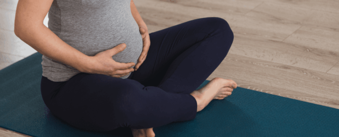 A pregnant is sitting cross-legged on a yoga mat.