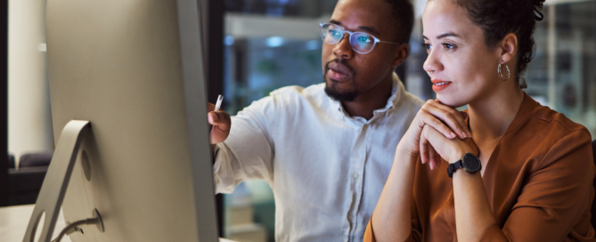 Man and a woman are sitting at a desk in an office setting, looking at a computer screen.