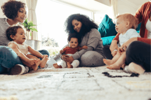 Three women and three infants, all seated on a rug in a living room.