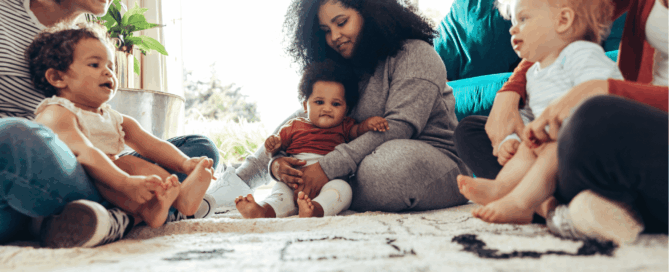 Three women and three infants, all seated on a rug in a living room.