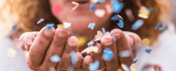 A person with warm, curly hair leans forward and gently blows a small storm of confetti from cupped hands.