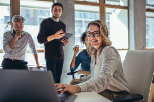 A woman with brown hair and glasses working on laptop while having meeting with colleagues in office.