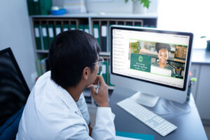 A man in a white lab coat is sitting at a desk, looking at a computer screen.