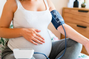 A pregnant woman resting one hand on her belly while the other arm is using a blood pressure monitor.