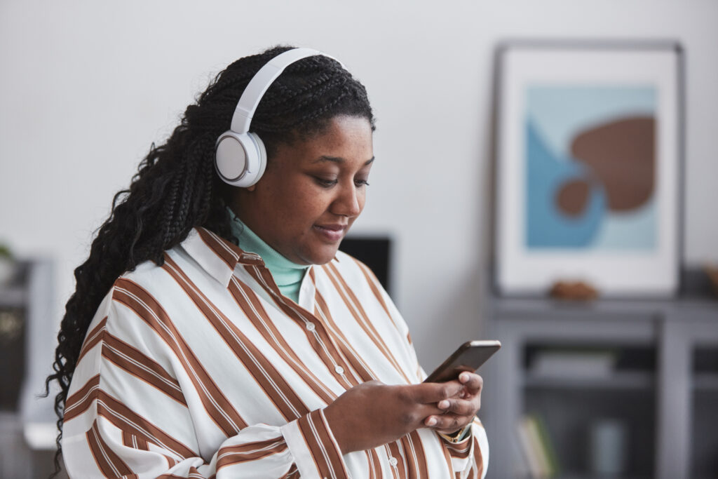 A young woman with long, curly hair is wearing white headphones and looking at her smartphone.