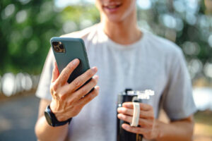 A person wearing a light gray t-shirt is holding a smartphone in one hand and a drink container in the other.