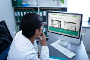 A person in a lab coat is sitting at a desk, looking at a computer screen.