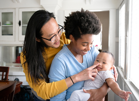 The man gently pokes the baby’s nose, while the woman looks on affectionately. A man and woman smiling as they interact with a baby.